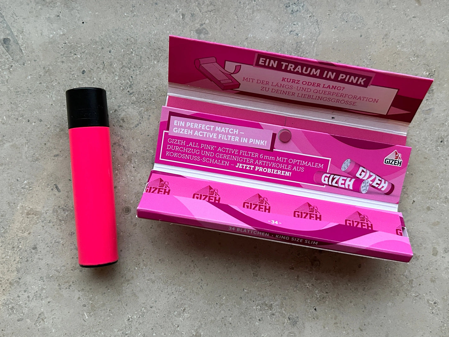 Ein rosa Clipper-Feuerzeug liegt neben einem offenen Ladys Starterpack von smokefy, das rosa Gizeh-Zigarettenfilter enthält, auf einer Marmoroberfläche. Das Paket ist mit mehreren Spitzen gefüllt und zeigt einen deutschen Text, der für das Produkt wirbt.